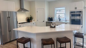 A modern kitchen with white cabinetry, a central island with stools, stainless steel appliances, and a window with a view. - Silver Lake & Dunes Dream House (Mears)