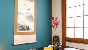 Een traditionele Japanse kamer met tatami vloerbedekking, een houten tafel gedekt met borden en stokjes, een hangende schrool met een berglandschap, en een vaas met rode bloemen.