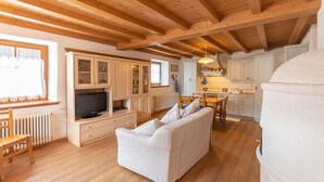 A spacious living area with a sofa, wooden ceiling beams, a television, and a kitchen with white cabinets. - Casa Roberto (Moena)