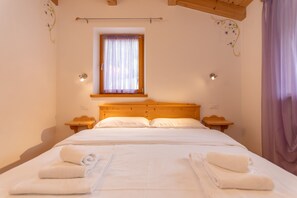 A bedroom with a wooden headboard, a window with curtains, two bedside tables, and a bed with neatly folded towels. - Villa Edelweiss Cardo (Alleghe)