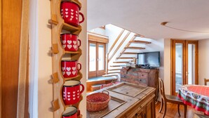 A kitchen with wooden shelves holding red mugs, a wooden dining table with chairs, and a staircase leading to another room.