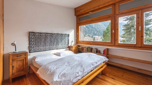 A bedroom with a wooden bed, a nightstand, a wall tapestry, and a window with a mountain view.