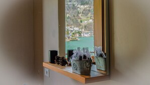 A window with a view of a body of water and hills, a wooden shelf with a pine cone, and two metal containers with items. - Regina 9 (Alleghe)