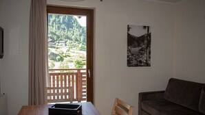 A room with a wooden table, a dark brown sofa, a framed black and white photograph on the wall, and a view of a mountainous landscape through a window. - Regina 14 (Alleghe)