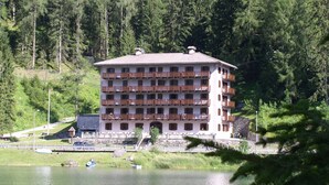 A multi-story hotel with balconies, situated by a lake surrounded by forest. - Regina 20 (Alleghe)
