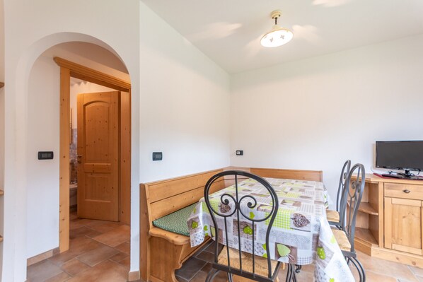 Une petite pièce accueillante, avec un banc en bois, une table recouverte d’une nappe à motifs, un téléviseur posé sur un meuble en bois et une porte donnant sur une autre pièce.