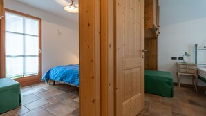 A bedroom with a bed, a wooden wardrobe, a green bench, a bedside table, and a window. - Casa De La Veisc (Pozza di Fassa)