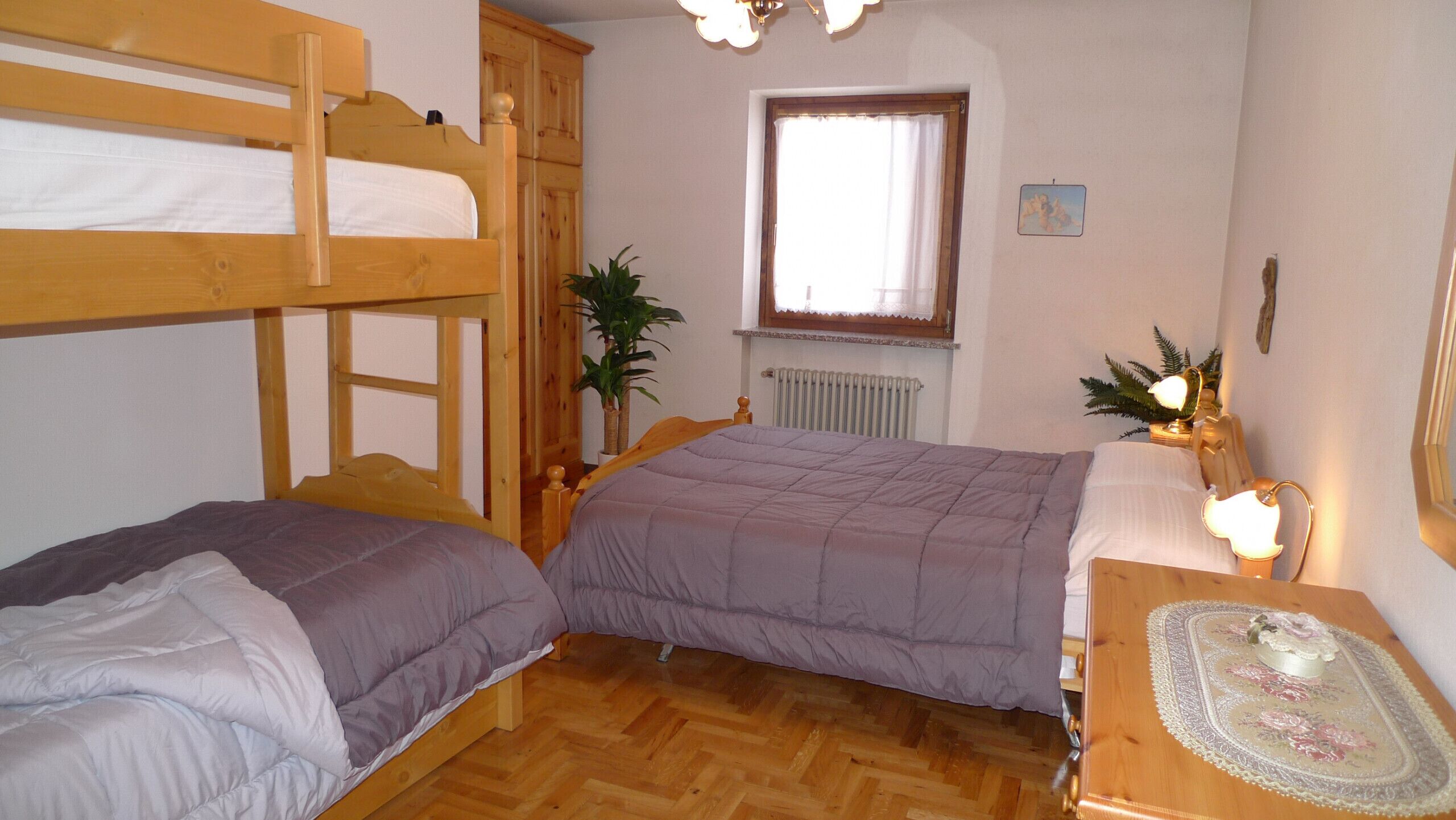 A room with two bunk beds, a window with a curtain, a radiator, and a small table with a lamp and decorative items.