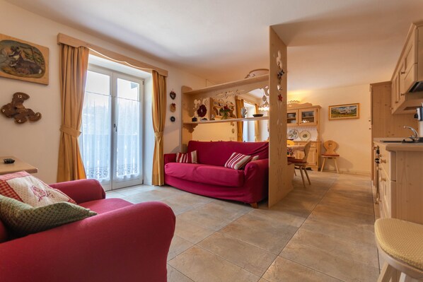 A living room with a red sofa, a dining area with a wooden table, and a kitchen with wooden cabinets. - Casa Rosi (Pozza di Fassa)