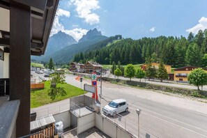 A view from a balcony overlooking a road with a car passing by, a parking sign, and a mountain range in the background. - Casa Rosi (Pozza di Fassa)