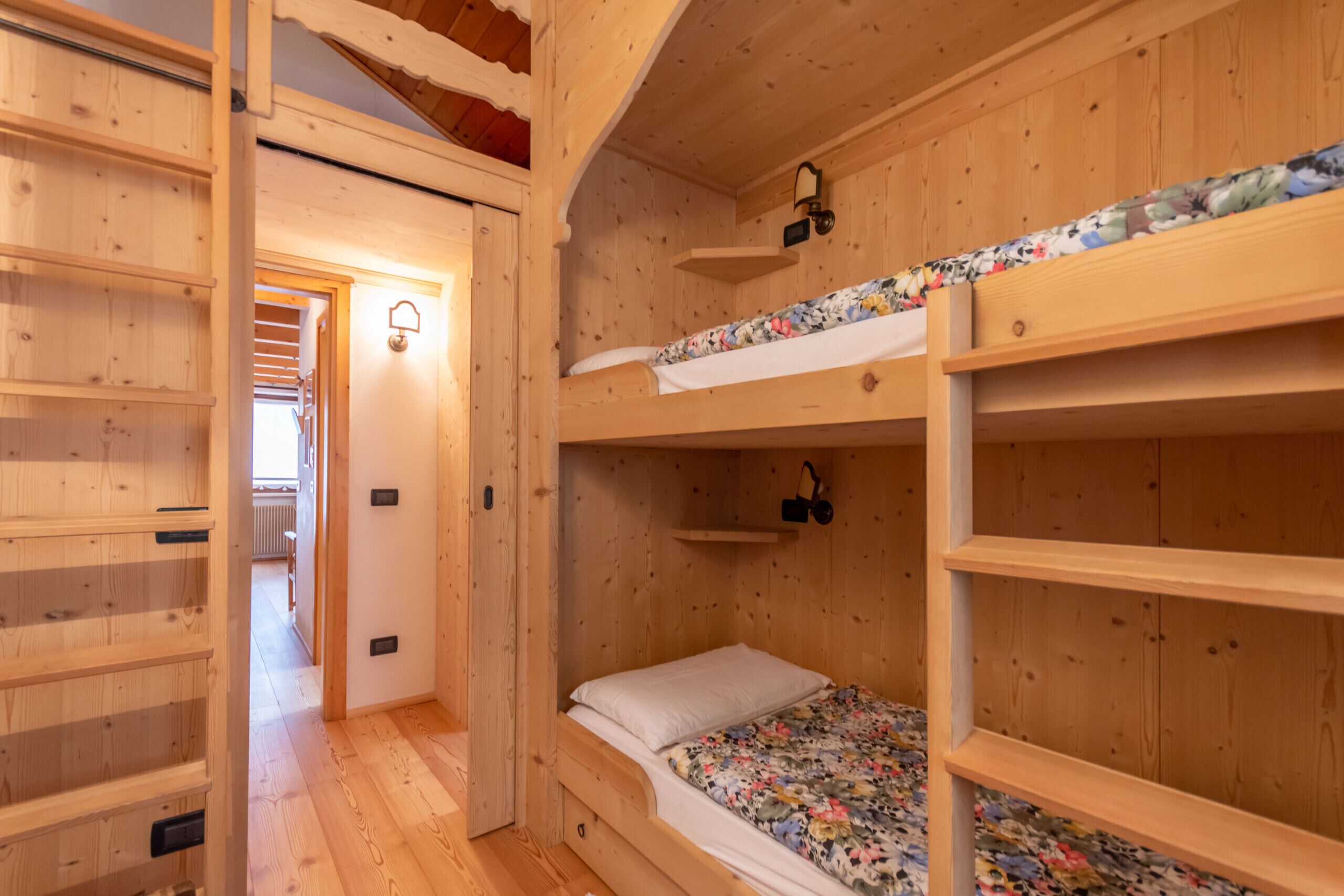 A wooden bunk bed room with a floral bedspread, a shelf above the bed, and a door leading to another room.