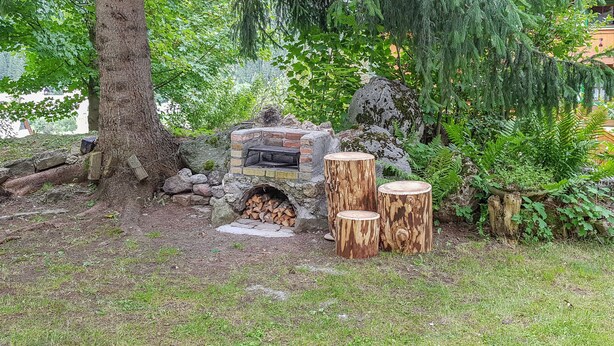 Une cheminée extérieure en pierre, entourée d’un décor naturel comprenant un arbre, des bûches et des tabourets en bois.