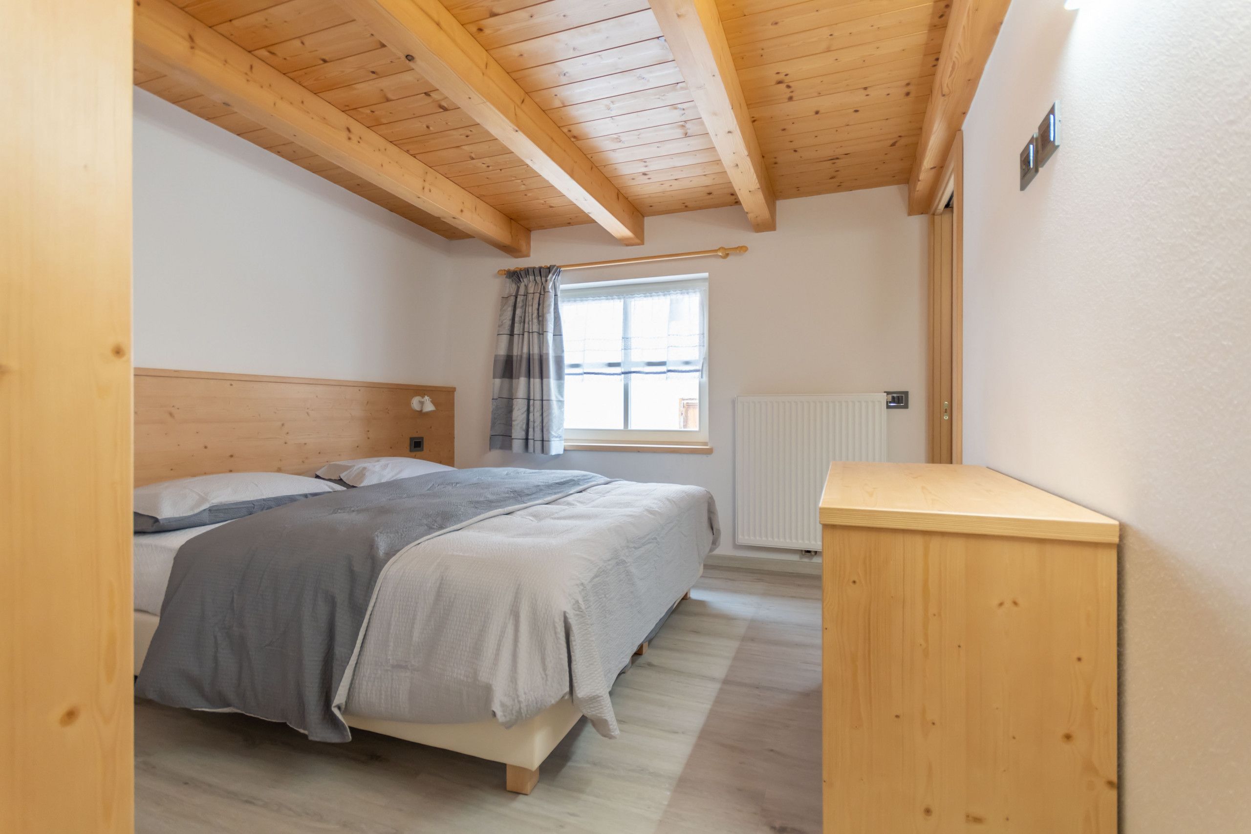 Une chambre avec un plafond en bois, un lit avec une literie grise, une fenêtre avec des rideaux et un radiateur.