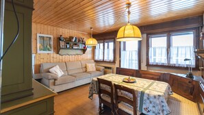 A cozy room with a wooden ceiling, a sofa, a dining table with a tablecloth, and a window with curtains.