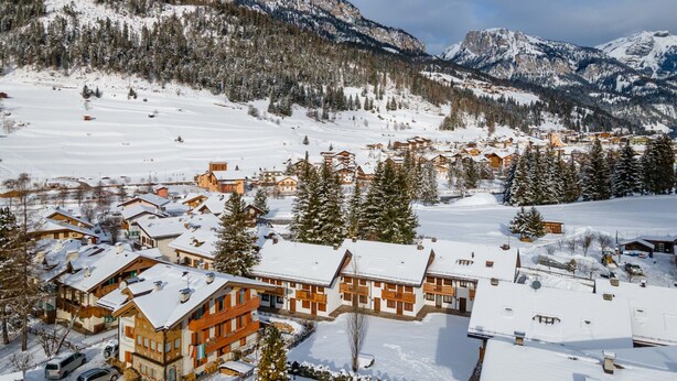 A snow-covered mountain village with chalet-style buildings, a ski slope, and a prominent mountain peak in the background.