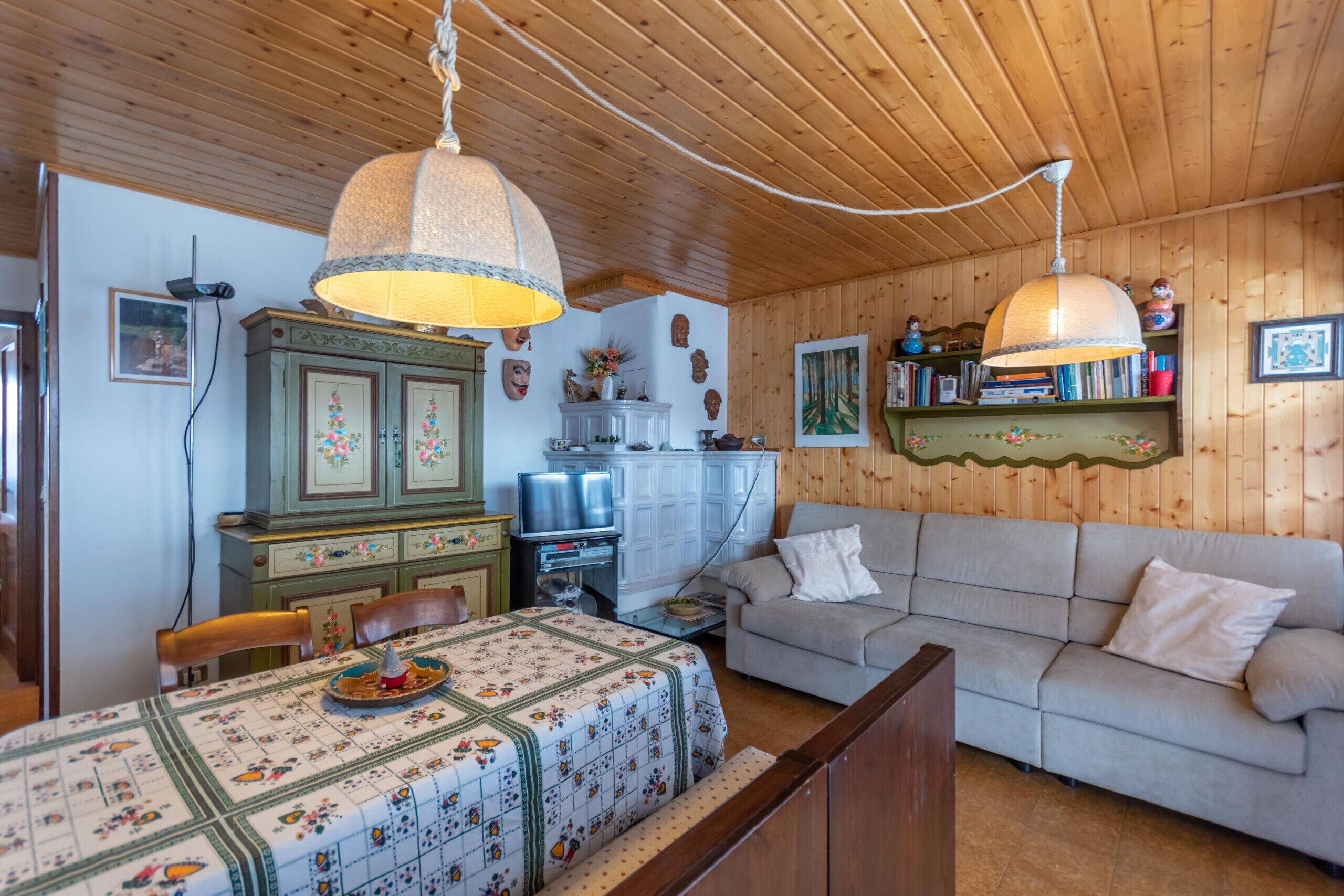 Ein gemütliches Wohnzimmer mit Holzwänden und -decke, einer Couch, einem Esstisch mit Blumenmuster-Tischdecke und einem Fernseher.