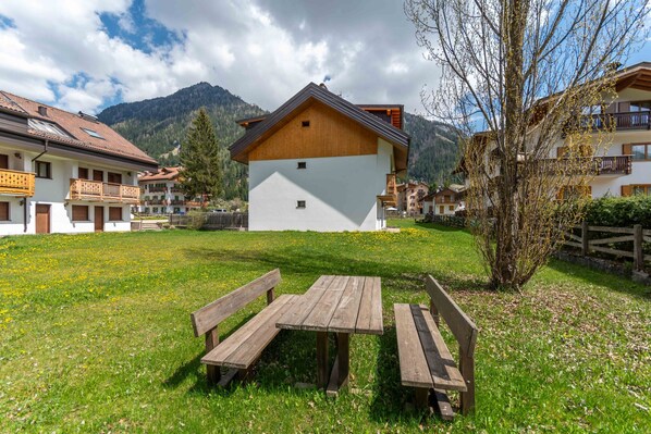 A building with a wooden facade and a white upper section, surrounded by a grassy area with benches. - Dimora al Vajolet (Pozza di Fassa)