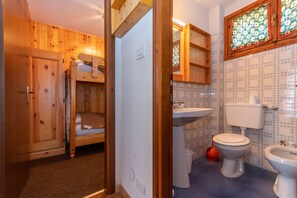 A wooden cabin-style room with bunk beds, a sink, toilet, and a window with patterned curtains.