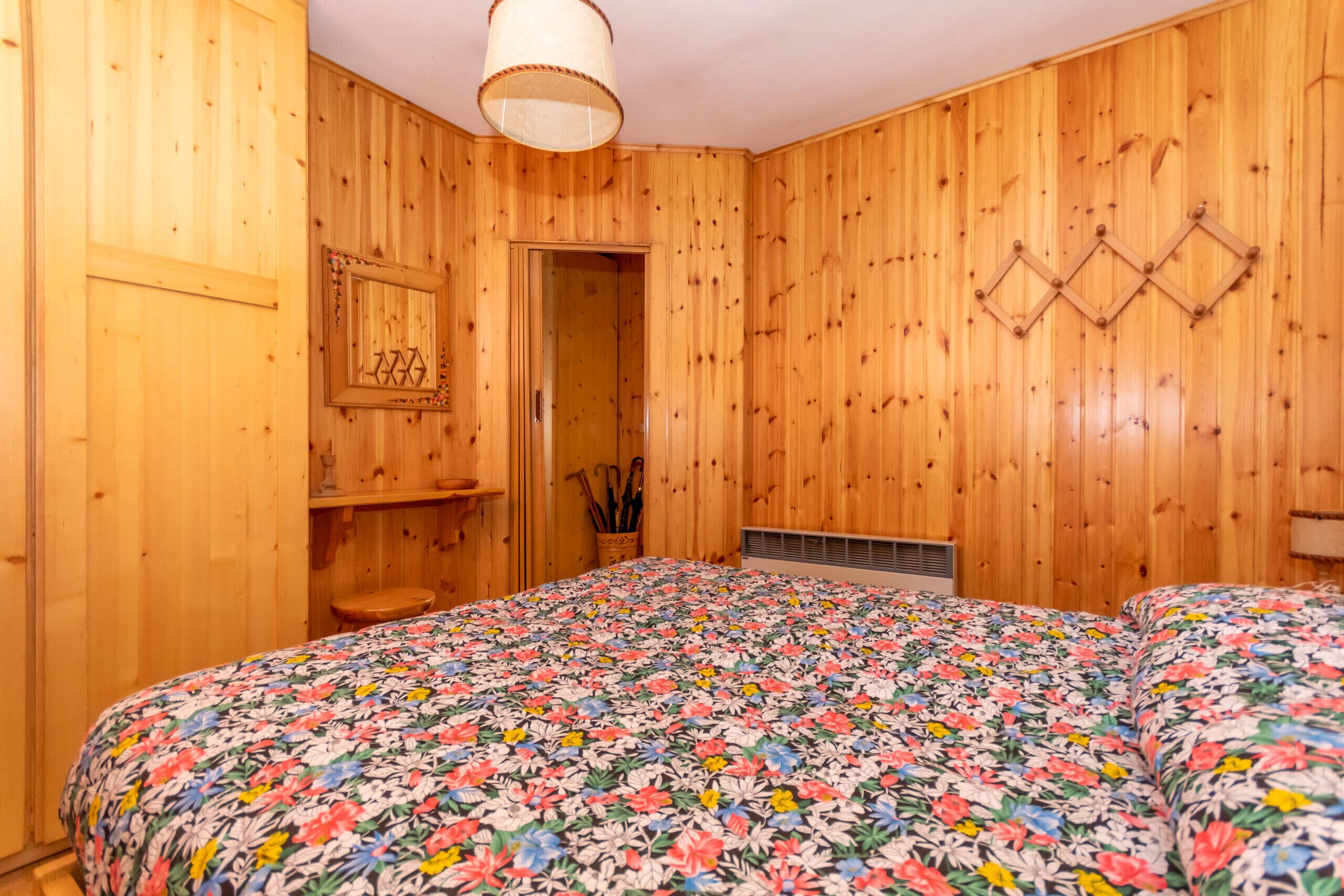 A wooden-paneled room with a bed, a door, a shelf with decorative items, and a wall-mounted light fixture.