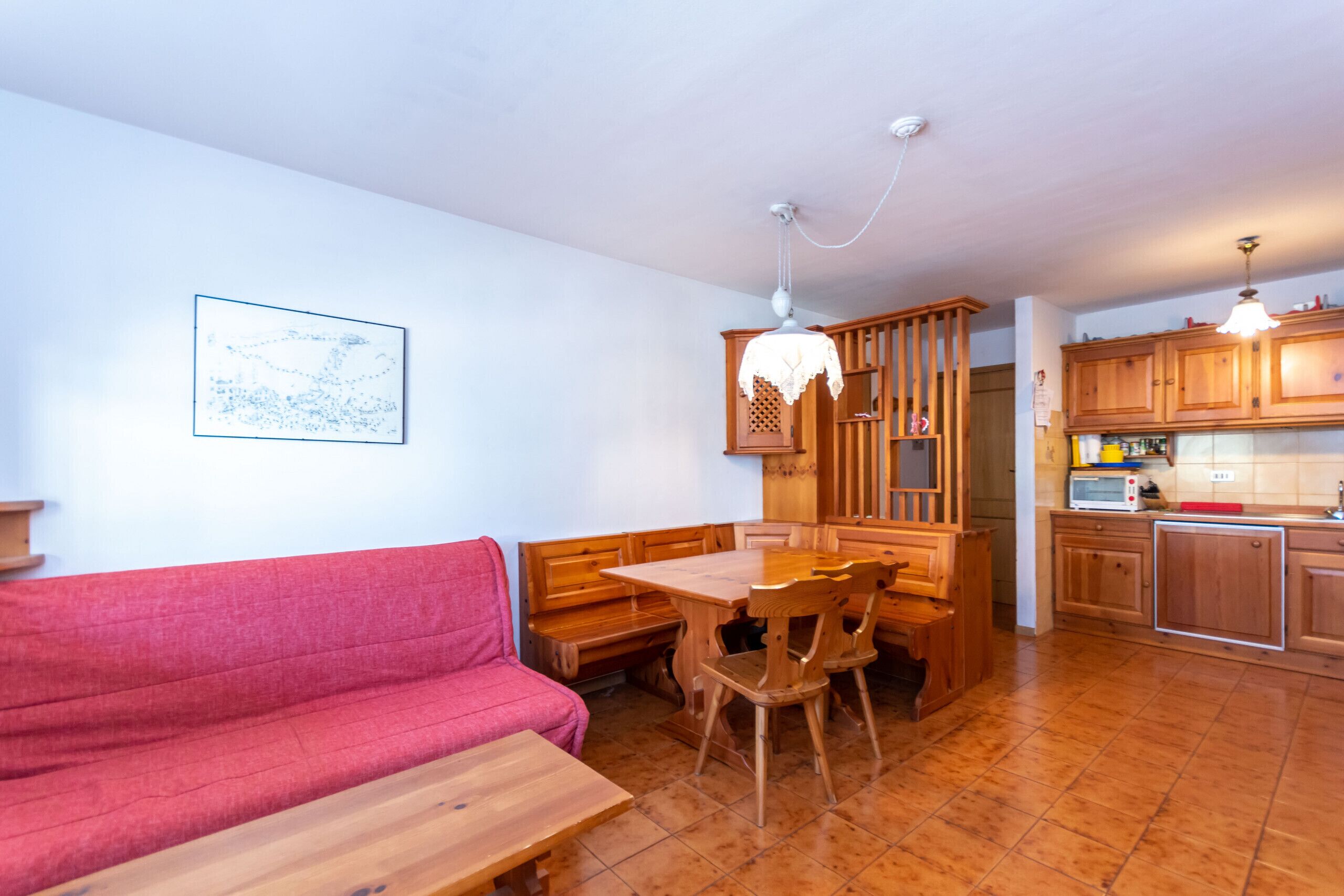 A cozy living area with a red sofa, wooden dining table, and chairs. A kitchen with wooden cabinets and a microwave is visible in the background.