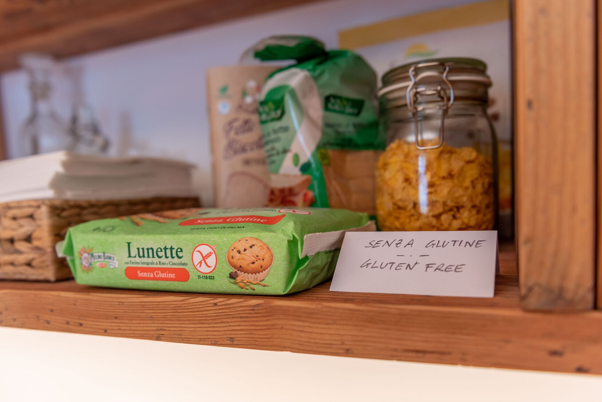 Una estantería con una caja de muffins sin gluten, un tarro de granola y una nota que dice 'Senza Glutine - Gluten Free'.