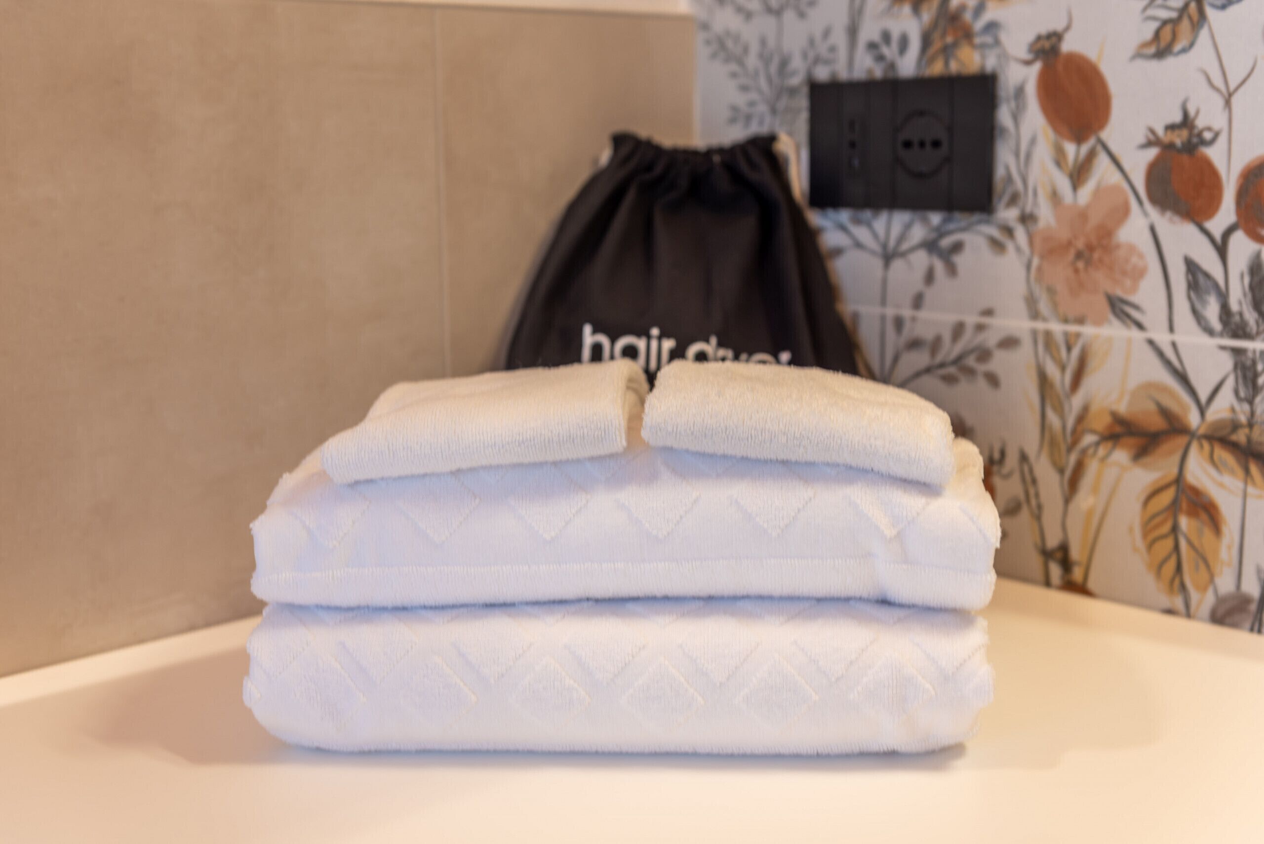 Une pile de serviettes blanches pliées est posée sur le comptoir d’une salle de bain dont le papier peint est à motifs floraux. Un sac noir à cordon, portant l’inscription « sèche-cheveux », se trouve à proximité.