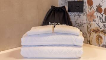 Une pile de serviettes blanches pliées est posée sur le comptoir d’une salle de bain dont le papier peint est à motifs floraux. Un sac noir à cordon, portant l’inscription « sèche-cheveux », se trouve à proximité.