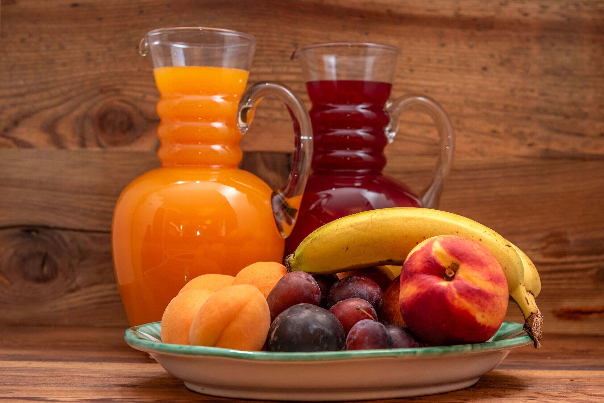 A wooden table with a glass pitcher of orange juice, a glass pitcher of red juice, a bowl of fruit including peaches, plums, a banana, and a peach.