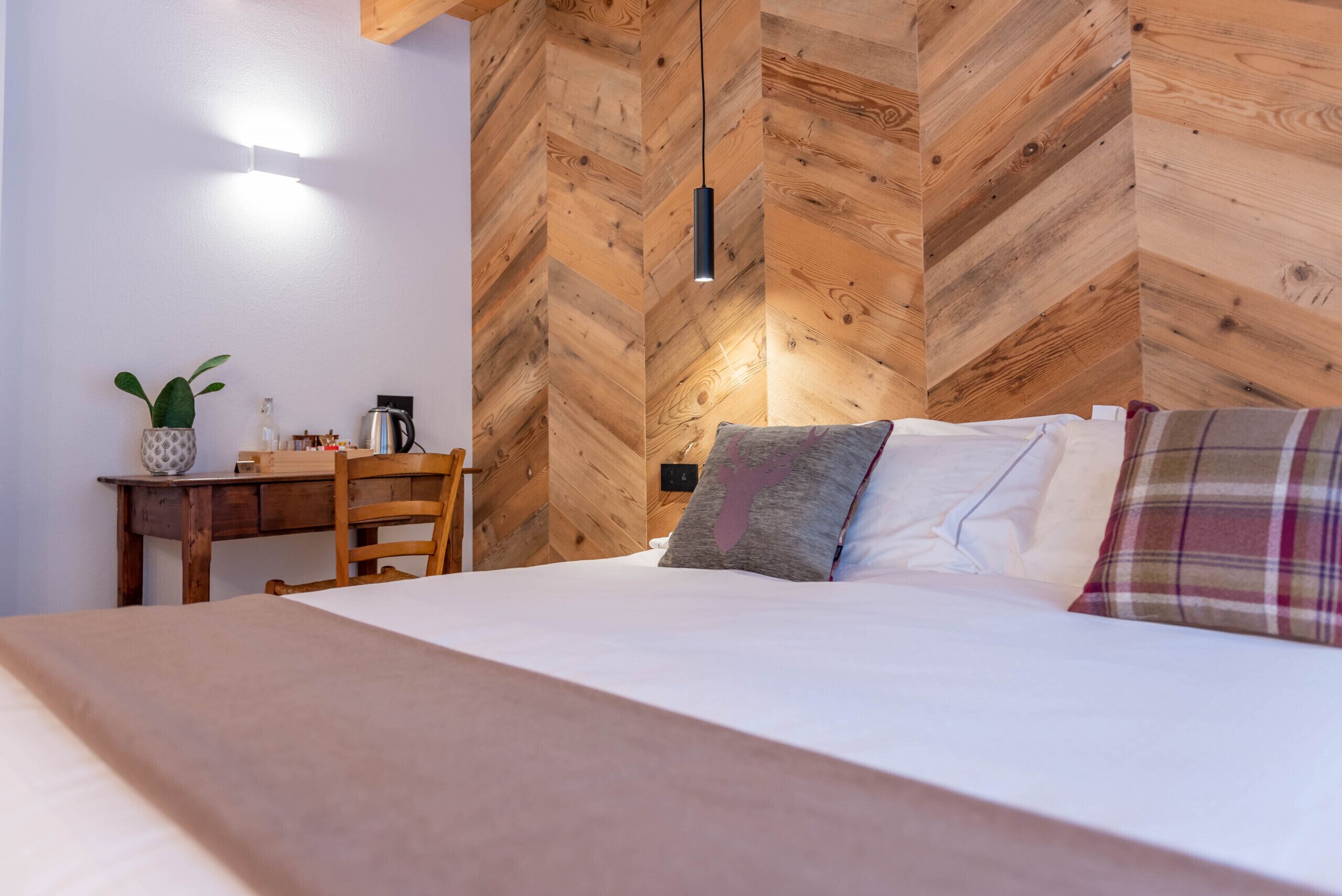Una camera da letto con parete in legno a spina di pesce, un letto con biancheria bianca, una scrivania con una pianta e una lampada a sospensione.