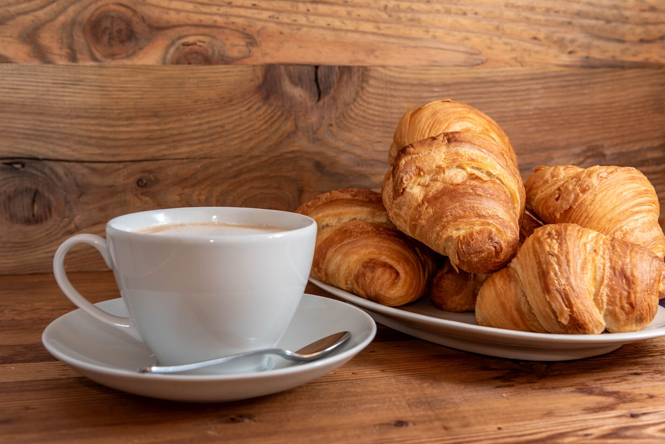 Eine weiße Tasse Kaffee auf einem Untertasse mit einem Löffel, neben einem Teller mit Croissants auf einem Holztisch.