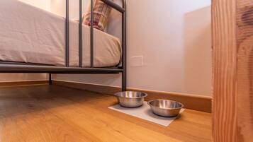 A small, neatly made bed with metal headboard and footboard, two metal bowls on a wooden floor mat, and a wooden door frame.