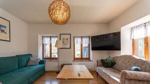 A living room with a wooden table, two sofas, a TV mounted on the wall, and a large window with curtains. - Casa Narciso (Rocca Pietore)