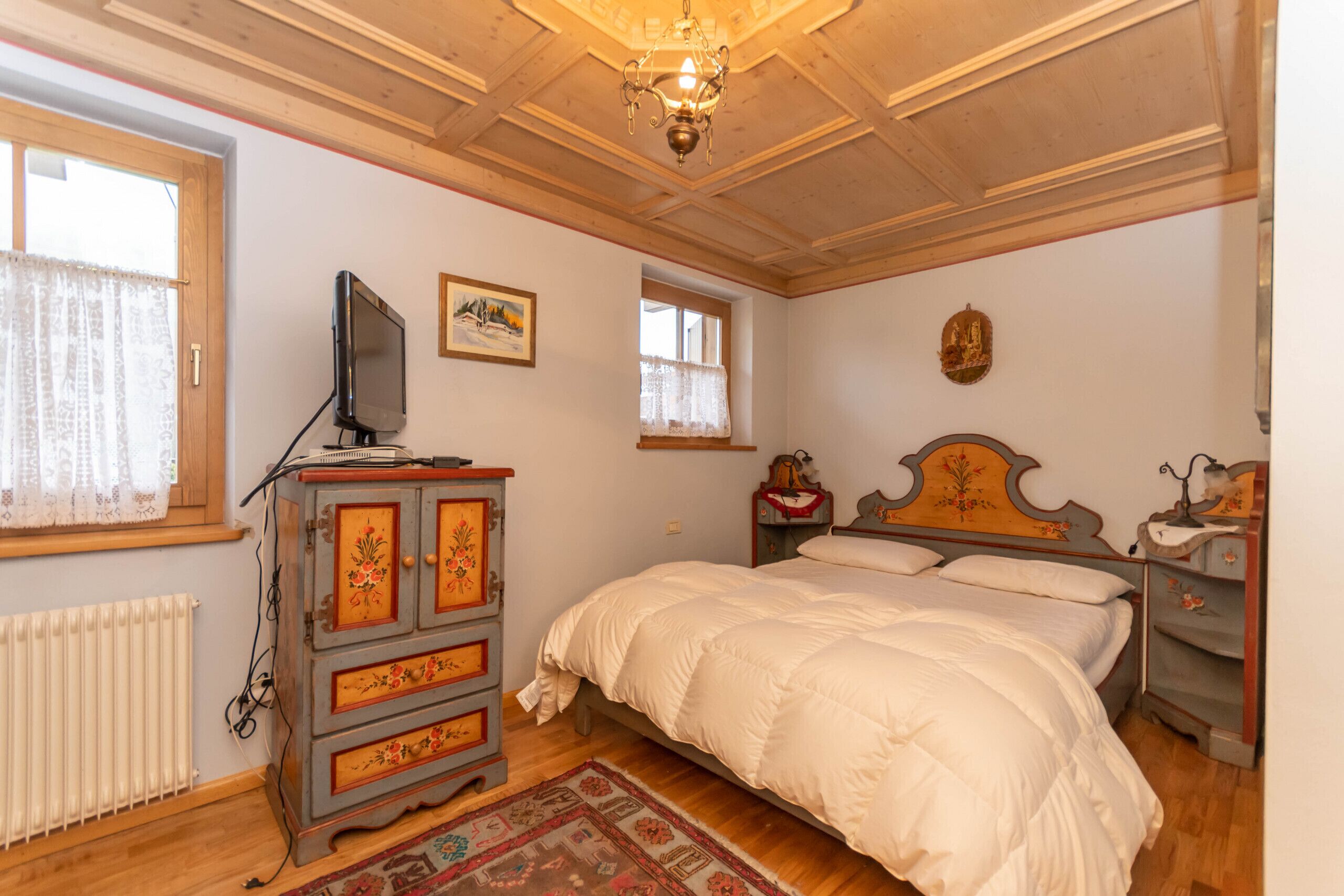 Een slaapkamer met een houten plafond, een bed met wit beddengoed, een dressoir met bloemenmotieven, een televisie op het dressoir en een raam met gordijn.