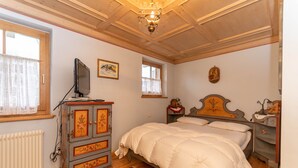 A bedroom with a wooden ceiling, a bed with white bedding, a dresser with floral designs, a television on the dresser, and a window with a curtain. - Casa Soreje (Mazzin)