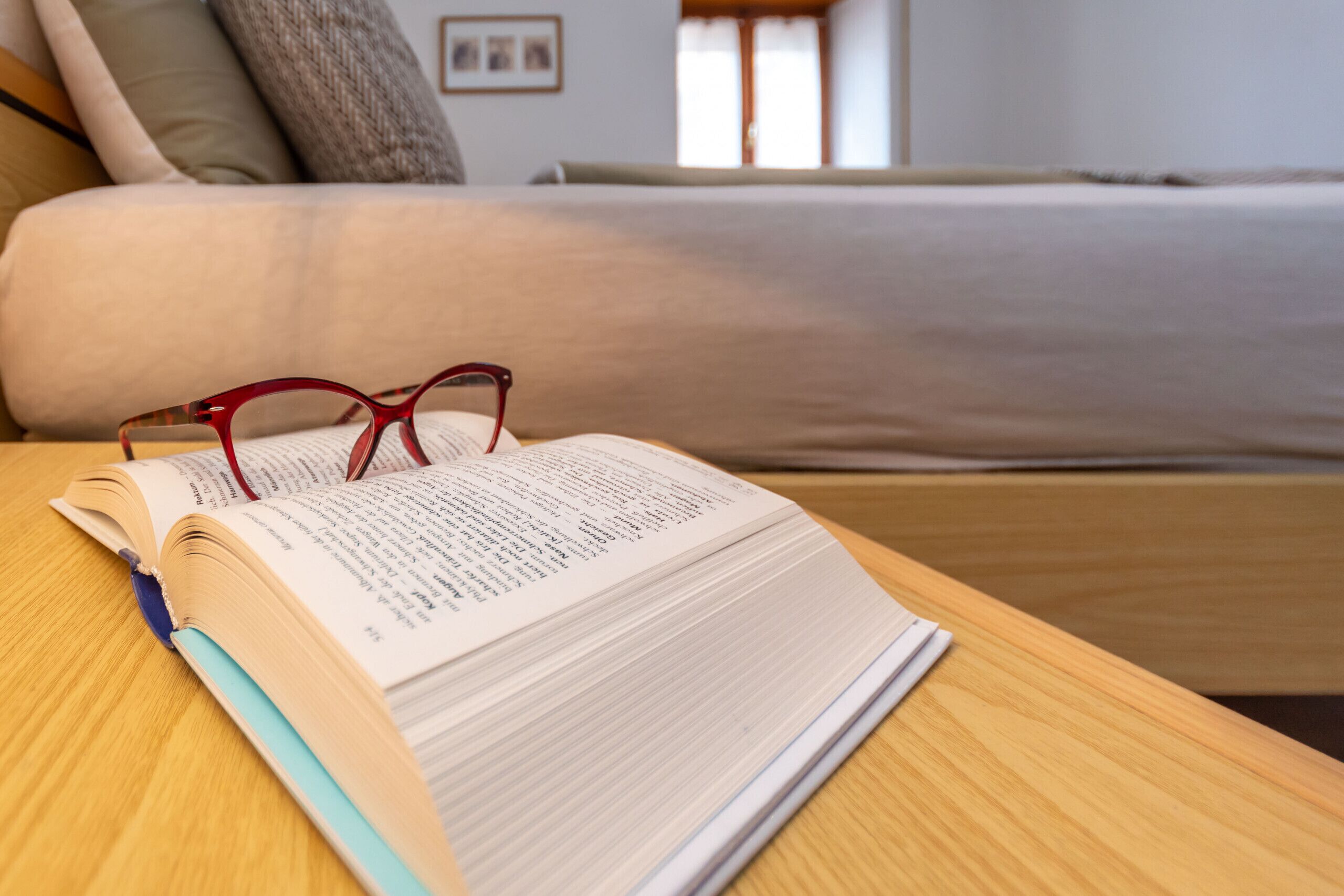An open book on a wooden table with a bed in the background.