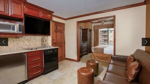 A compact hotel room with a kitchenette, a brown leather sofa, and a view of a building through an open door.