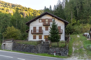 Een chalet met twee verdiepingen, met een stenen trap die naar de ingang leidt, omgeven door een houten omheining en een beboste helling.