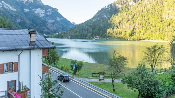 A house with a solar panel roof, a road with a car, and a lake surrounded by mountains. - mansarda Lungolago (Alleghe)