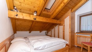 A wooden bed with white bedding in a room with a sloped ceiling and a small window with lace curtains. - mansarda Lungolago (Alleghe)
