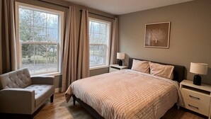 Un dormitorio con una cama grande, un sofá, dos mesitas de noche con lámparas y una ventana con cortinas.