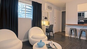 Ein modernes Wohnzimmer mit einem weißen Sofa, einem runden Couchtisch mit einer Zeitschrift, einem Schreibtisch mit Lampe und einer Küchenzeile inklusive Mikrowelle und Hockern.