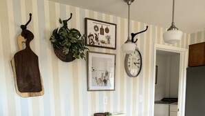 A kitchen with striped wallpaper, a wooden cutting board, a plant, framed pictures, a clock, and a modern bar stool setup.
