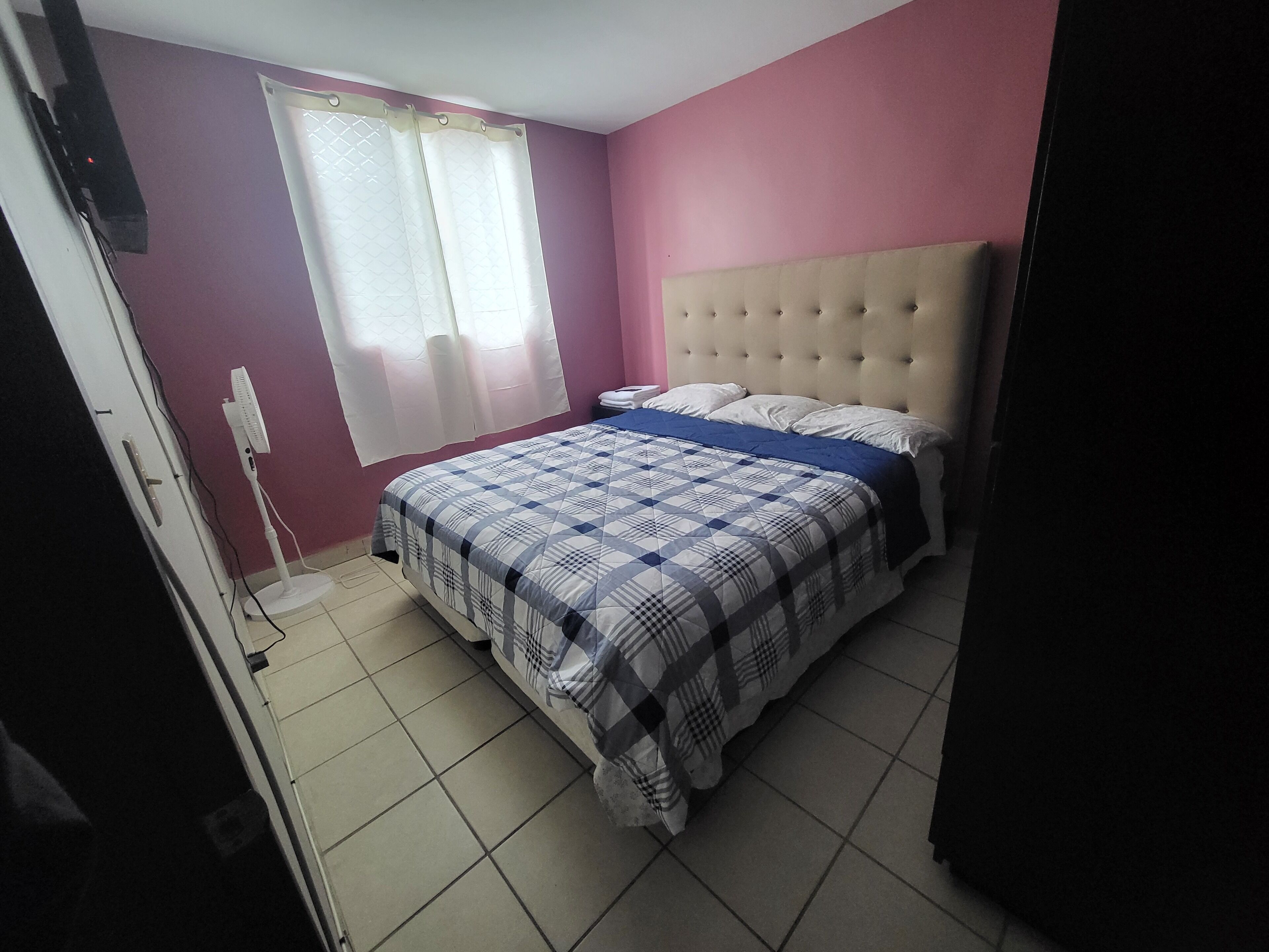 A bedroom with a bed, a window with curtains, a bedside table, and a wall-mounted TV.