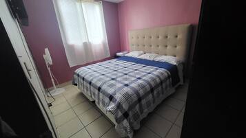 A bedroom with a bed, a window with curtains, a bedside table, and a wall-mounted TV.