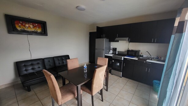 A compact living space with a dining table, chairs, a black sofa, a kitchenette, and a ceiling-mounted security camera. - Departamentos velasco (Tijuana)