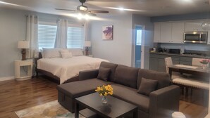 A modern living room with a sofa, a coffee table with a vase, a bed with a nightstand, a ceiling fan, and a kitchen area with cabinets and appliances. - STUDIO COMFORT (Stanton)