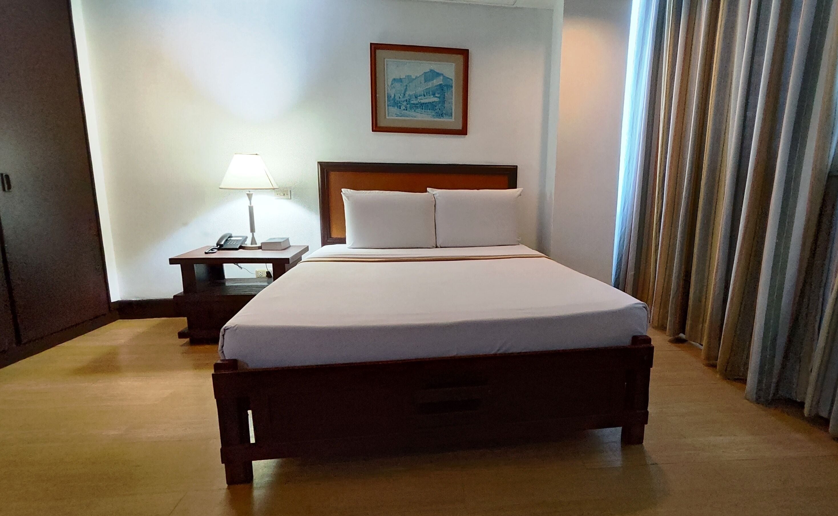 Habitación de hotel con cama, mesita de noche con lámpara, un cuadro enmarcado en la pared y una ventana con cortinas.