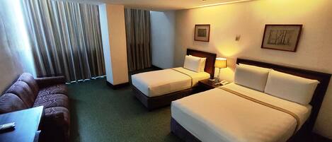 A hotel room with two single beds, a sofa, a desk, and a window with curtains.