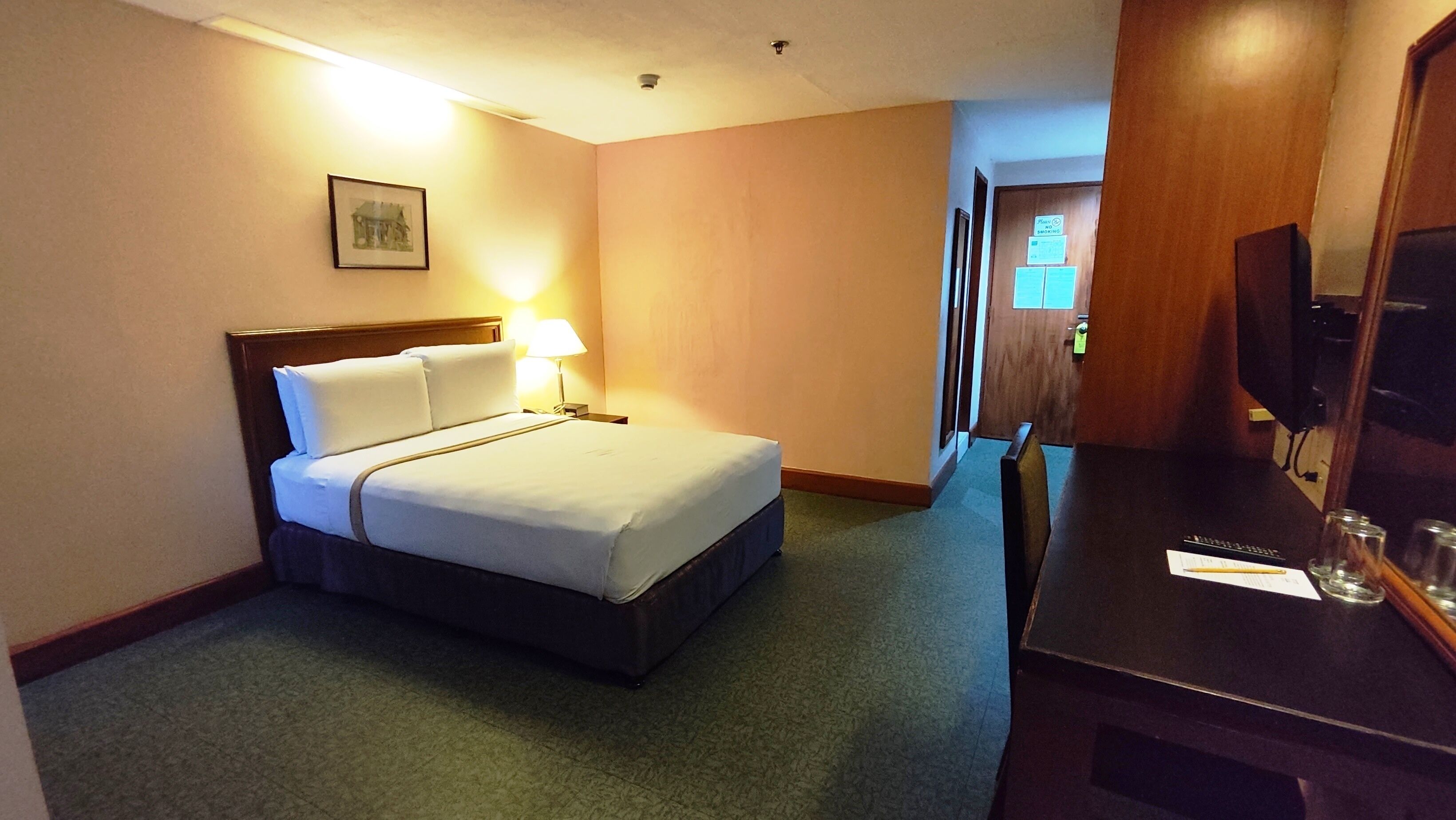 Una habitación de hotel con una cama, lámpara de noche, escritorio con silla y televisor.