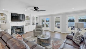 A spacious living room with a fireplace, comfortable seating, a television, and large windows offering a view of a lake.
